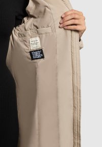 Bež prešita jakna z gladko teksturo, ki vsebuje oznako za nego in blagovno znamko znotraj. Zunanja tkanina se zdi lahka in mehka.