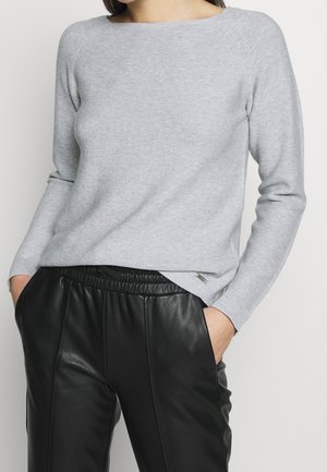Femme portant un pull gris clair à manches longues et un pantalon en cuir noir, les mains dans les poches, sur un fond uni.