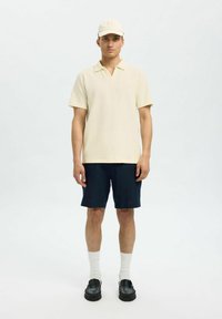 Polo tričko s krátkým rukávem textured ve světlé žluté barvě, kombinované s námořnickými šortkami. Bílá ponožky a černé mokasíny dotvářejí outfit.