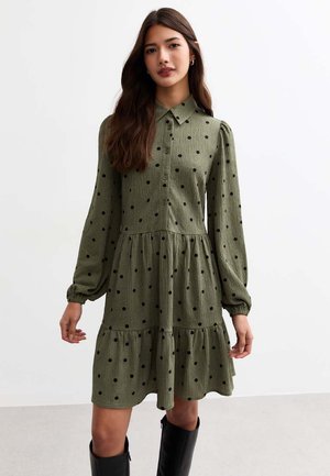Robe verte en tissu texturé, dotée d'un col, de longues manches bouffantes et de pois noirs. Détail d'ourlet en niveaux.