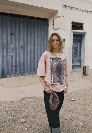 Jasnoróżowa koszula z krótkim rękawem z nadrukiem graficznym i akcentami geometrycznymi. Czarne luźne spodnie i szare kalosze z gumy uzupełniają strój.