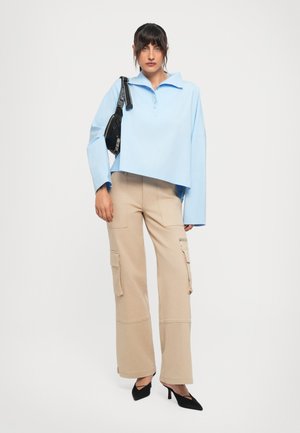 Donna che indossa una camicia oversize azzurro chiaro, pantaloni cargo beige, tacchi neri e porta una borsa a tracolla nera su uno sfondo bianco puro.