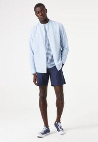 Lichtblauwe overhemd met lange mouwen over een marineblauw gestreept T-shirt, gecombineerd met marineblauwe shorts en blauwe sneakers. Beschikt over een knoopsluiting.