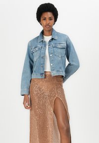 Denimska jakna v svetlo modri barvi z gumbnim zapiranjem, prsni žepi in dolgimi rokavi, v kombinaciji z bronasto bleščečo krilom z razporkom ob strani.
