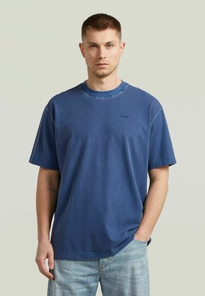 Junger Mann mit kurzen Haaren trägt ein blaues, übergroßes T-Shirt und hellblaue Jeans und steht vor einem schlichten hellen Hintergrund.