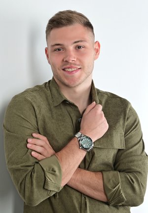 Green textured shirt, silver watch with green dial. The shirt has a button-up collar and rolled sleeves, arms crossed in front.