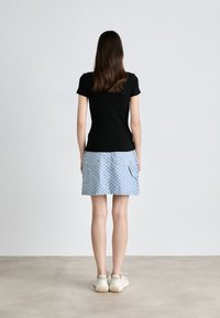Femme debout, dos à la caméra, portant un t-shirt noir, une jupe à carreaux bleu clair avec poches et des baskets blanches sur fond uni.