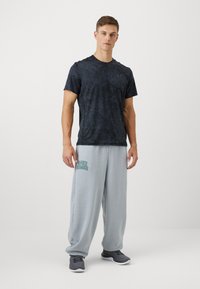 Černé tričko s krátkým rukávem s motivem, nošené s světle šedými tepláky s logem "Under Armour" v tyrkysové barvě. Šedé sportovní boty dotvářejí celý vzhled.
