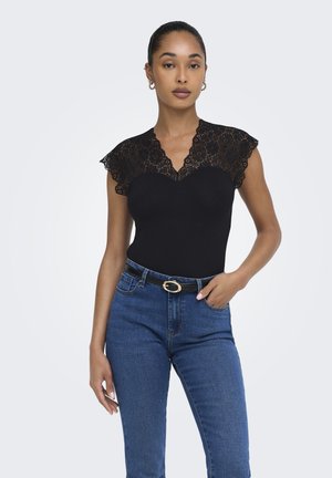 Femme portant un haut en dentelle noire à manches courtes, rentré dans un jean bleu taille haute avec une ceinture noire, debout avec une main dans la poche devant un fond uni.