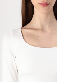 Balta ilgomis rankovėmis palaidinė su apvaliu kaklu, prigludusia siluetu ir lygiu, tamprių audinio tekstūra. Minimalūs siūlės ir jokie matomi raštai.