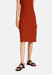 Sandali neri con motivo floreale, suola piatta e due cinghie, indossati con un vestito aderente, lungo fino al ginocchio, di colore ruggine uniforme.