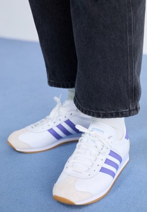 Baskets blanches avec des rayures violettes portées avec des chaussettes blanches et un pantalon noir sur un sol bleu clair.