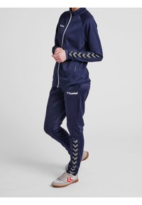 Marinträningsdress i slät tyg; zip-up jacka med chevronmönster på ärmarna, matchande byxor med sidodetaljer och grå skor.