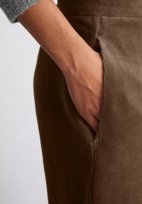 Brauner Cord-Rock mit vertikaler Ripp-Textur, ausgestattet mit einer Seitentasche und einer taillierten Taille. Ein Arm in einem hellgrauen Strickpullover ist sichtbar.