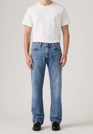 Homme portant un t-shirt blanc uni à manches courtes, un jean bleu clair droit et des mocassins noirs, debout devant un fond uni.