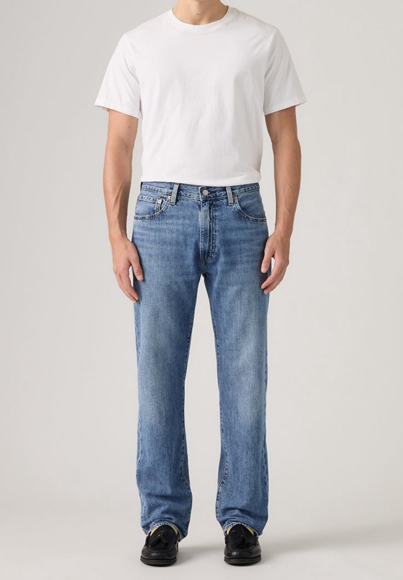 Homme portant un t-shirt blanc uni à manches courtes, un jean bleu clair droit et des mocassins noirs, debout devant un fond uni.