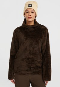 Brun, fyldig fleece-pullover med høj krave og lange ærmer, der har en afslappet pasform. Båret med en beige beanie-hat med logo.