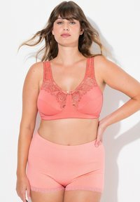 Koraalroze bh met kantdetails en brede bandjes, gecombineerd met bijpassende polkadotshorts. Zacht materiaal en textuurcontrast van kant zijn opvallend.