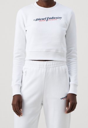 Person iført en hvid cropped sweatshirt og matchende hvide sweatpants, begge med "Diesel Industry Denim Division" logo i blåt og rødt.
