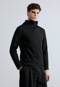 Sudadera negra con cremallera, fabricada en tela suave, con capucha, bolsillos delanteros y un logo discreto en el pecho. Combinada con pantalones joggers negros a juego.