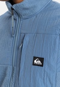Fleecová bunda světle modré barvy s látkou v mřížkovém vzoru, vysokým límcem a zipovým zapínáním. Obsahuje boční kapsu a nášivku s logem Quiksilver.