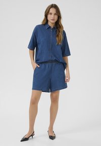 Denim kaheosaline komplekt, mis sisaldab lühikeste varrukatega nööpidega särki ja elastse vöökohtadega lühikesi pükse. Mõlemad on keskmiselt sinises toonis kontrastsete õmblustega.