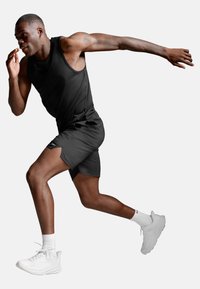 Atletisk mand i sort tanktop og shorts, der sprintet, iført hvide sokker og løbesko, isoleret på lys baggrund.