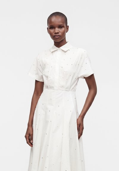 Mannequin féminin portant une robe blanche à manches courtes avec un col et de petites décorations éparpillées sur le tissu, debout devant un fond uni.
