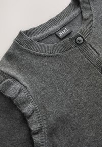 Cardigan gris en maille avec encolure ronde côtelée, fermeture par un bouton noir unique et détail volanté le long de la couture de l’épaule.