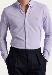 Mężczyzna w jasnofioletowej koszuli zapinanej na guziki, włożonej w spodnie o wysokim stanii w czarne paski, z rękami w kieszeniach.