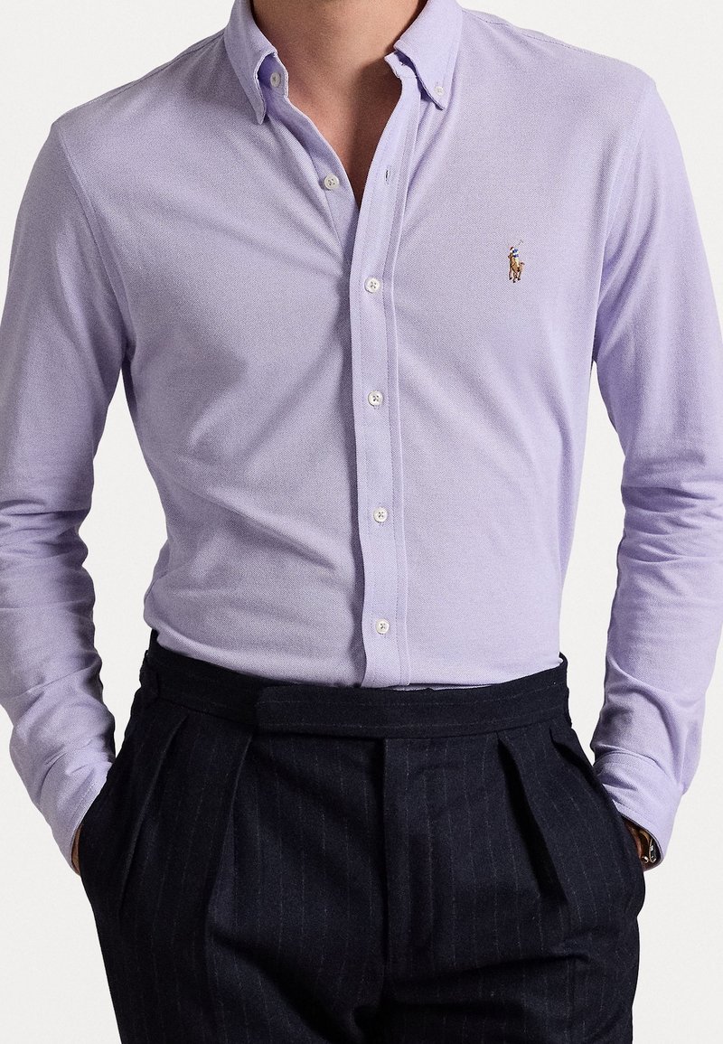 Mężczyzna w jasnofioletowej koszuli zapinanej na guziki, włożonej w spodnie o wysokim stanii w czarne paski, z rękami w kieszeniach.