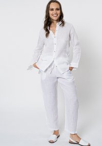 Camicia bianca di lino con bottoni e maniche lunghe, abbinata a pantaloni di lino coordinati. La modella indossa sandali bianchi. Tessuto liscio e leggero.