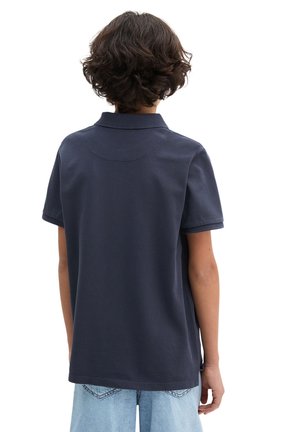 Kind mit lockigen braunen Haaren, trägt ein marineblaues kurzärmeliges Hemd und hellblaue Jeans, steht mit dem Rücken zum Betrachter vor einem weißen Hintergrund.