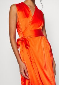 Pomarańczowa sukienka kopertowa z satynowym wykończeniem, głębokim dekoltem w kształcie litery V i wiązanym paskiem w talii. Materiał jest gładki, o subtelnym połysku i elegancko się układa.