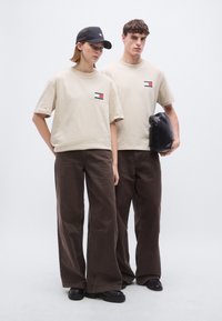 Μπεζ βαμβακερά t-shirts με μικρό λογότυπο, συνδυασμένα με σκούρα καφέ παντελόνια φαρδιάς γραμμής. Μία μαύρη καπέλο και τσάντα συμπληρώνουν τις εμφανίσεις.