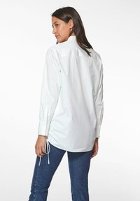 Weißes Langarmhemd mit spitzem Kragen, gerafften Seiten und geknöpften Manschetten, kombiniert mit dunkelblauen Jeans. Glatte Textur, lässiges Design.