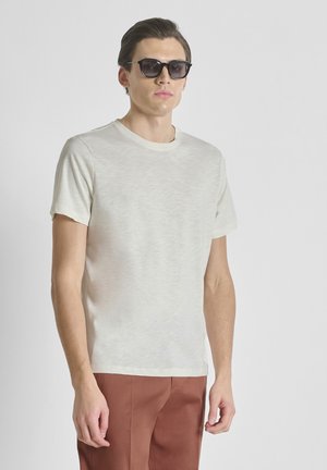 Uomo con occhiali da sole rettangolari neri, maglietta a maniche corte color avorio e pantaloni color ruggine su uno sfondo semplice.