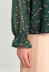 Blouse à manches longues florale en tissu transparent vert foncé, ornée de fleurs rose clair et poignets élastiqués, associée à une jupe plissée beige.