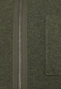 Närbild av olivgrön stickad tyg med en central silverfärgad metalldragkedja och en liten rektangulär patchficka på högra sidan.