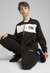 Czarna i biała kurtka PUMA z guzikami, z logo na piersi. Model nosi dopasowane czarne dresy i ciemne sneakersy.