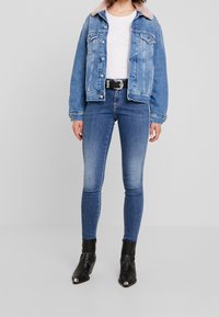 Giacca di jeans con un lavaggio azzurro chiaro, caratterizzata da un colletto di pelliccia rosa, due tasche sul petto e bottoni argentati, abbinata a jeans skinny blu scuro e stivaletti neri.