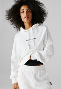 Jonge vrouw met krullend haar, gekleed in een witte "Red Bull Racing" hoodie en bijpassende witte joggingbroek, voor een effen grijze achtergrond.