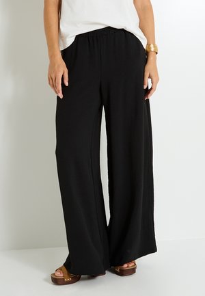 Pantalons larges noirs en tissu texturé avec une taille élastique. Associés à des sandales compensées marron, offrant une coupe décontractée.