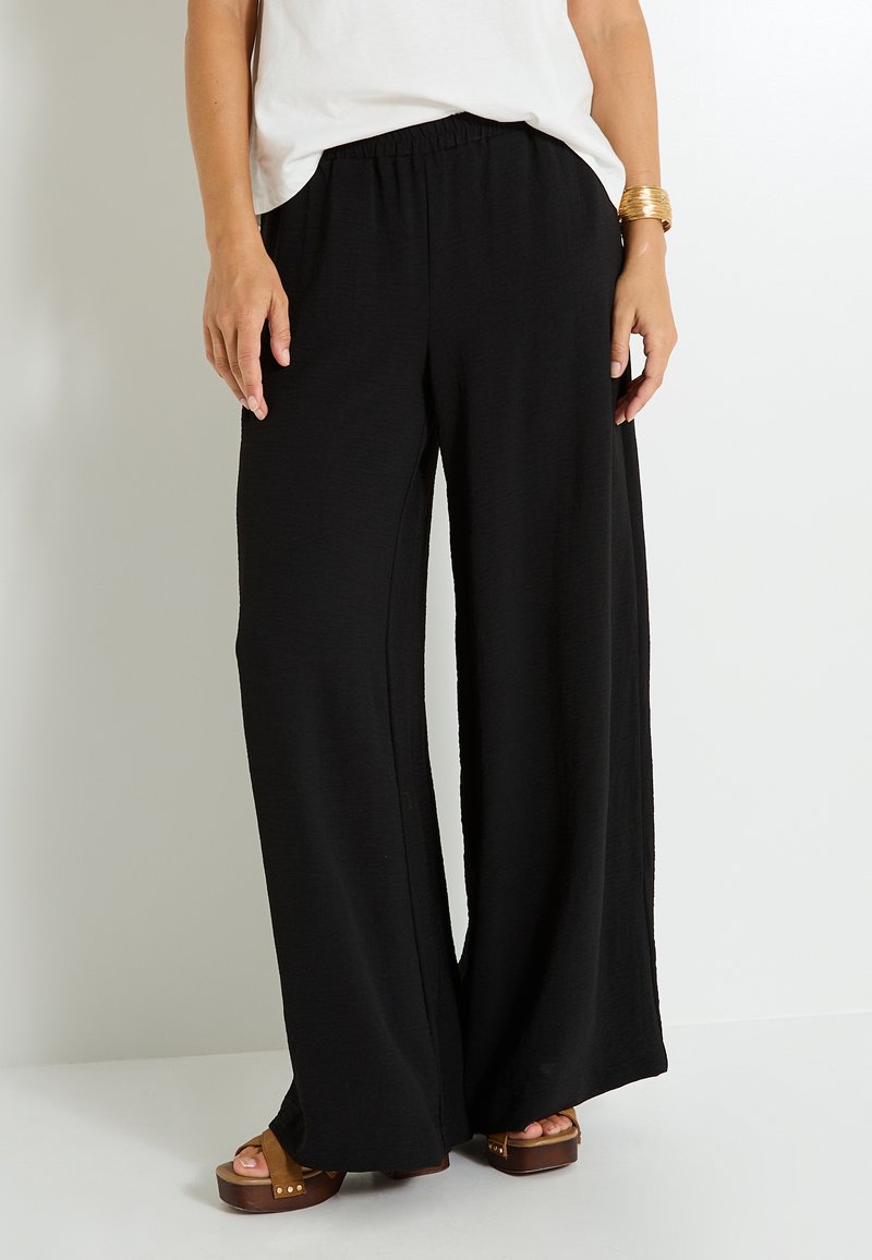 Pantalones de pierna ancha negros hechos de una tela texturizada con una cinturilla elástica. Combinados con sandalias de plataforma marrones, mostrando un ajuste relajado.