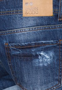 Jeans de mezclilla azul oscuro con un bolsillo trasero desgastado con bordes deshilachados, costuras naranjas en contraste y una etiqueta de marca de cuero marrón.