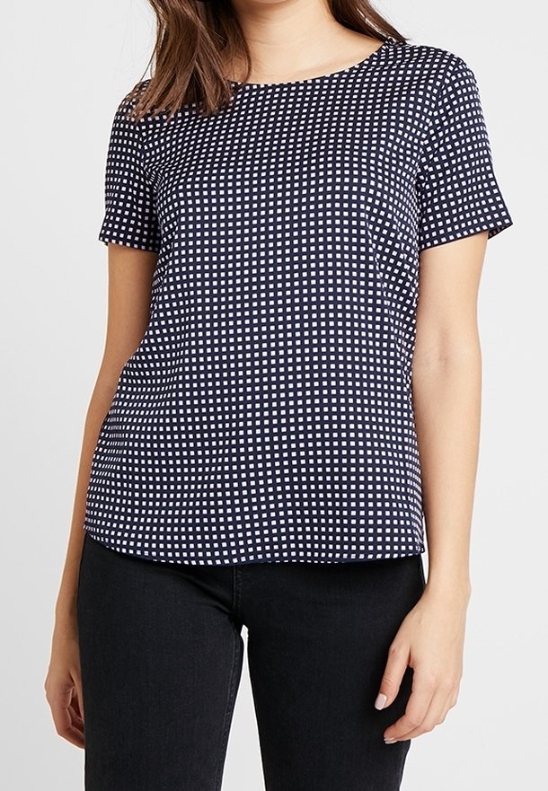 Femme portant un haut bleu marine à manches courtes avec un petit motif carré blanc et un pantalon noir, debout devant un fond uni.