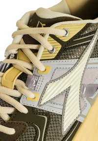 Sneaker con una toma superiore in brown e grigio testurizzato, con dettagli gialli e grigi, lacci color crema e un design a strati. Elementi riflettenti e lisci presenti.