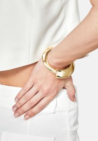 Bracciale rigido circolare color oro con una finitura liscia e riflettente e un accento appuntito. Indossato su un polso con abbigliamento bianco.