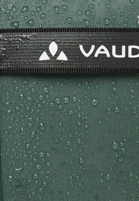 Groene stof met waterdruppels, voorzien van een zwarte band met het witte "Vaude" logo en driehoekige ontwerpelementen. Textuurdetails zijn zichtbaar.