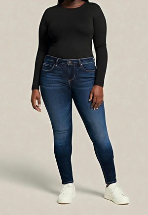 Osoba nosząca ciemnoniebieskie wąskie dżinsy, czarną bluzkę z długim rękawem i białe trampki, stojąca na jednolitym jasnym tle.